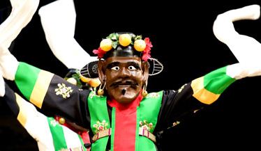 A dancer in a mask representing Cheoyong. The dark-faced mask smiles wide. Its rugged features include deep-set eyes, prominent nose and cheekbones, and facial hair.