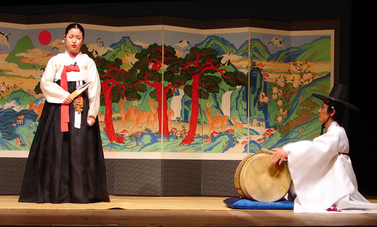A female-presenting person in traditional Korean hanbok holding a closed fan stands before a painted screen, with a male-presenting person in hanbok sitting off to the right playing a drum.